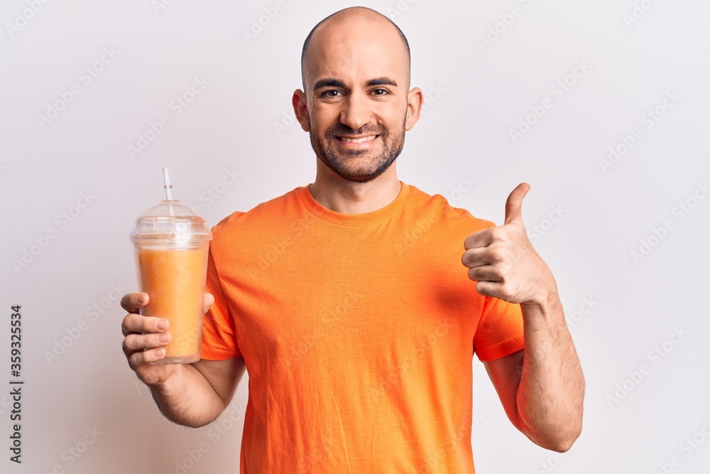 Young handsome bald man drinking glass of healthy orange juice smiling happy and positive, thumb up doing excellent and approval sign Young handsome bald man drinking glass of healthy orange juice smiling happy and positive, thumb up doing excellent and approval sign