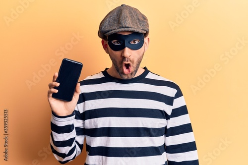 Young handsome bald man wearing burglar mask holding smartphone scared and amazed with open mouth for surprise, disbelief face © Krakenimages.com