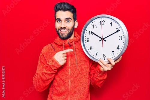 Young handsome man with beard holding big clock smiling happy pointing with hand and finger