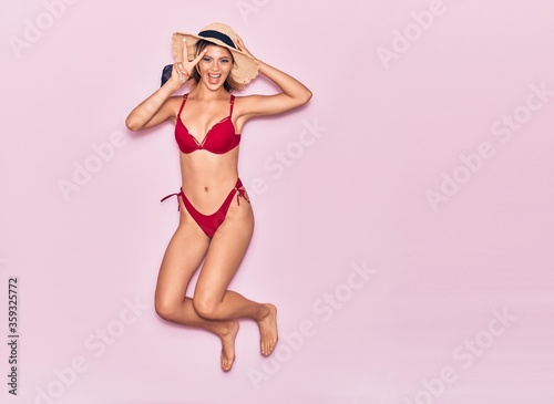 Young beautiful girl on vacation wearing bikini and summer hat smiling happy. Jumping with smile on face doing victory sign over isolated pink background