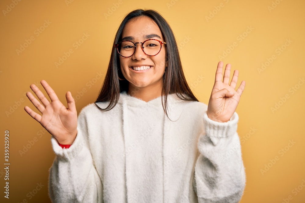 Young beautiful asian woman wearing casual sweater and glasses over yellow background showing and pointing up with fingers number nine while smiling confident and happy. Young beautiful asian woman wearing casual sweater and glasses over yellow background showing and pointing up with fingers number nine while smiling confident and happy.
