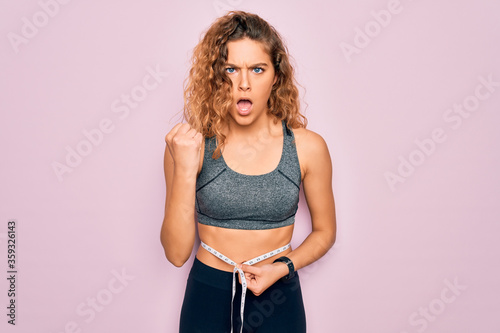 Young blonde sporty woman with blue eyes controlling weight using tape measure on waist annoyed and frustrated shouting with anger, crazy and yelling with raised hand, anger concept © Krakenimages.com