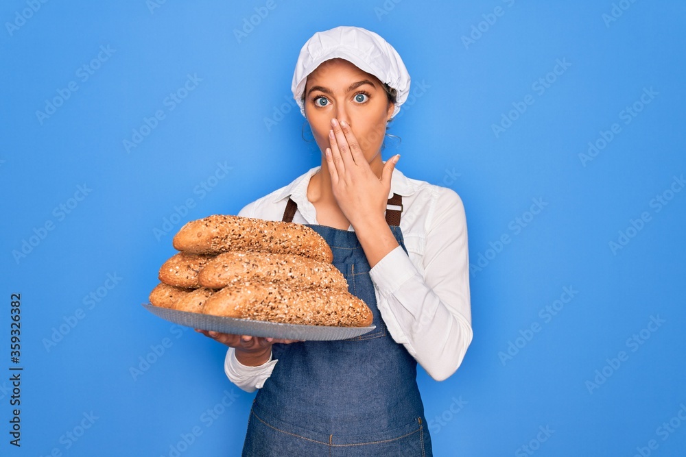 Young beautiful blonde baker woman with blue eyes holding tray with wholemeal bread cereal cover mouth with hand shocked with shame for mistake, expression of fear, scared in silence, secret concept Young beautiful blonde baker woman with blue eyes holding tray with wholemeal bread cereal cover mouth with hand shocked with shame for mistake, expression of fear, scared in silence, secret concept
