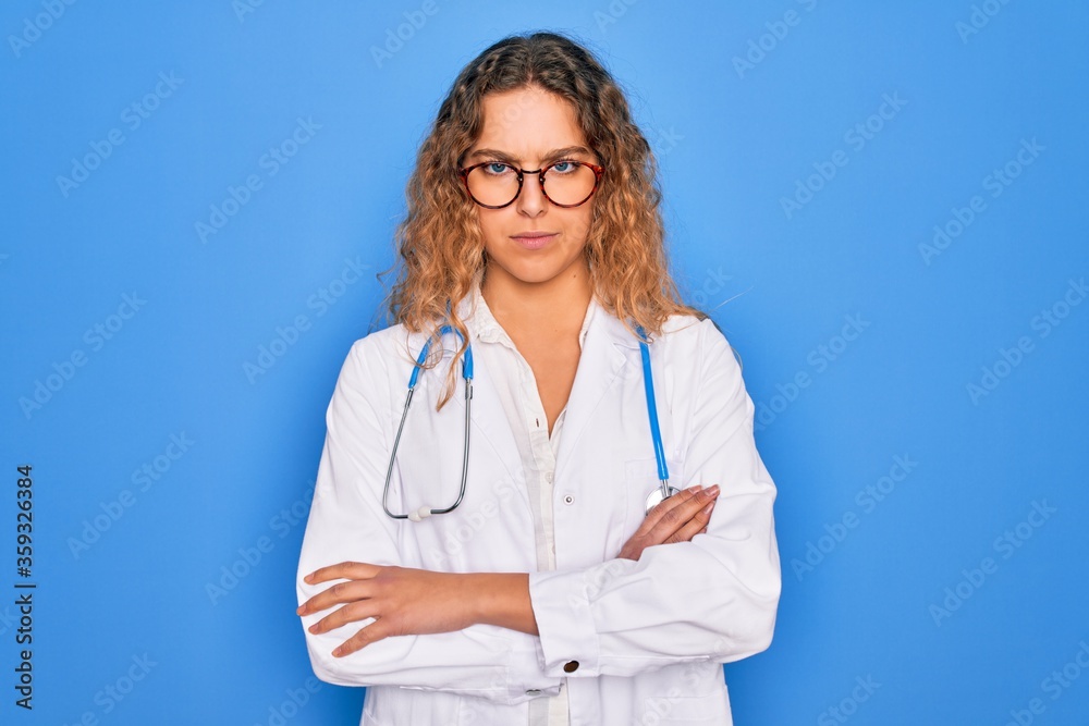 Young beautiful blonde doctor woman with blue eyes wearing coat and stethoscope skeptic and nervous, disapproving expression on face with crossed arms. Negative person. Young beautiful blonde doctor woman with blue eyes wearing coat and stethoscope skeptic and nervous, disapproving expression on face with crossed arms. Negative person.