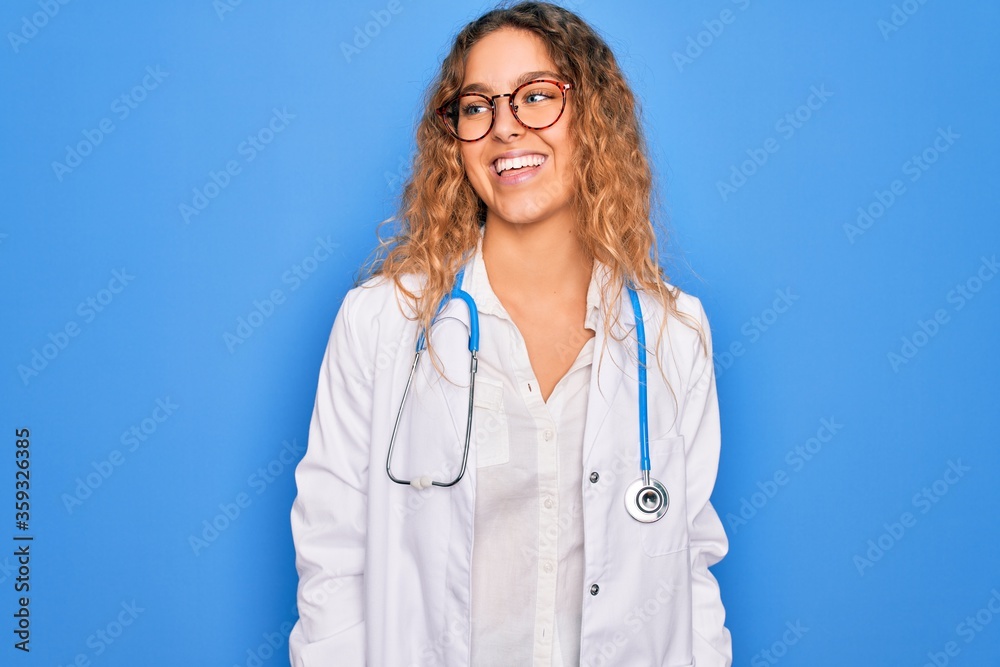 Young beautiful blonde doctor woman with blue eyes wearing coat and stethoscope looking away to side with smile on face, natural expression. Laughing confident. Young beautiful blonde doctor woman with blue eyes wearing coat and stethoscope looking away to side with smile on face, natural expression. Laughing confident.