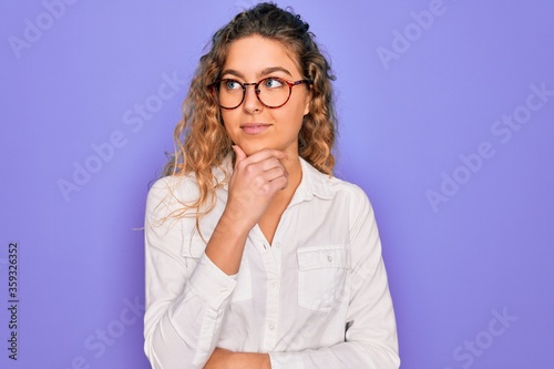 Young beautiful woman with blue eyes wearing casual shirt and glasses over purple background with hand on chin thinking about question, pensive expression. Smiling with thoughtful face. Doubt concept.