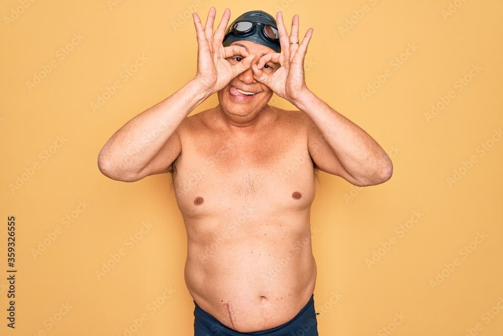 Middle age senior grey-haired swimmer man wearing swimsuit, cap and goggles doing ok gesture like binoculars sticking tongue out, eyes looking through fingers. Crazy expression.