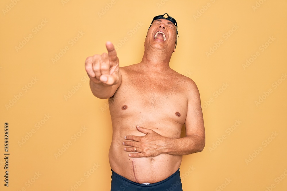 Middle age senior grey-haired swimmer man wearing swimsuit, cap and goggles laughing at you, pointing finger to the camera with hand over body, shame expression