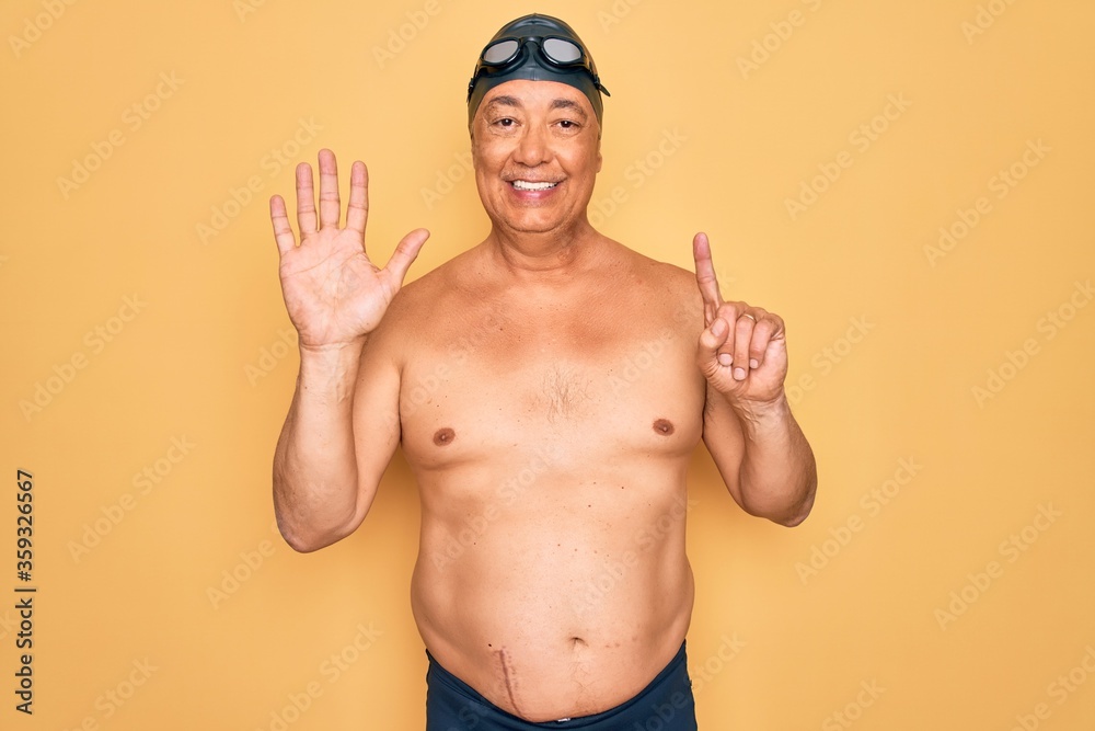 Middle age senior grey-haired swimmer man wearing swimsuit, cap and goggles showing and pointing up with fingers number six while smiling confident and happy.