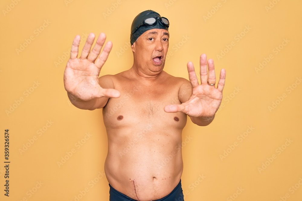 Middle age senior grey-haired swimmer man wearing swimsuit, cap and goggles doing stop gesture with hands palms, angry and frustration expression