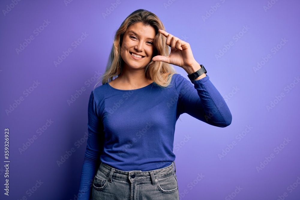 Young beautiful blonde woman wearing casual t-shirt over isolated purple background smiling and confident gesturing with hand doing small size sign with fingers looking and the camera. Measure Young beautiful blonde woman wearing casual t-shirt over isolated purple background smiling and confident gesturing with hand doing small size sign with fingers looking and the camera. Measure