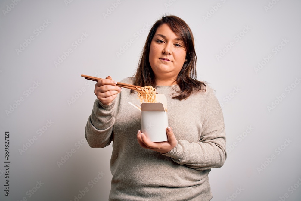 Beautiful plus size woman eating asian noodles food from box over isolated background with a confident expression on smart face thinking serious Beautiful plus size woman eating asian noodles food from box over isolated background with a confident expression on smart face thinking serious