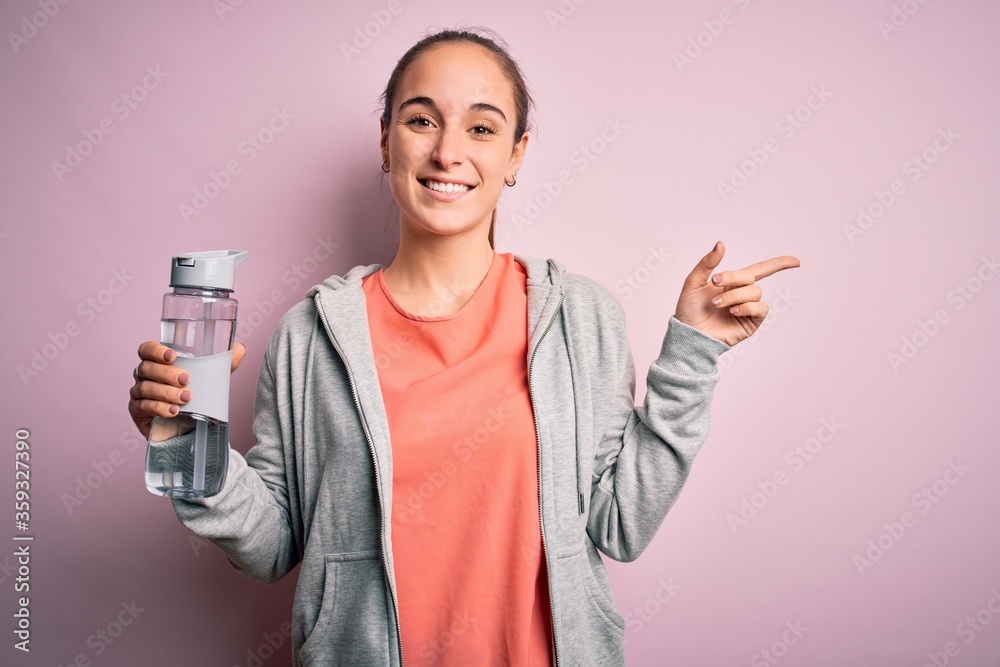 Young beautiful sporty woman doing sport drinking bottle with water to refreshment very happy pointing with hand and finger to the side Young beautiful sporty woman doing sport drinking bottle with water to refreshment very happy pointing with hand and finger to the side