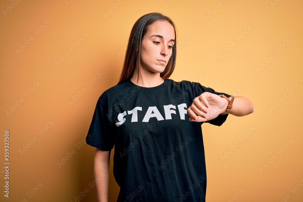 Young beautiful worker woman wearing staff uniform t-shirt over isolated yellow background Checking the time on wrist watch, relaxed and confident Young beautiful worker woman wearing staff uniform t-shirt over isolated yellow background Checking the time on wrist watch, relaxed and confident