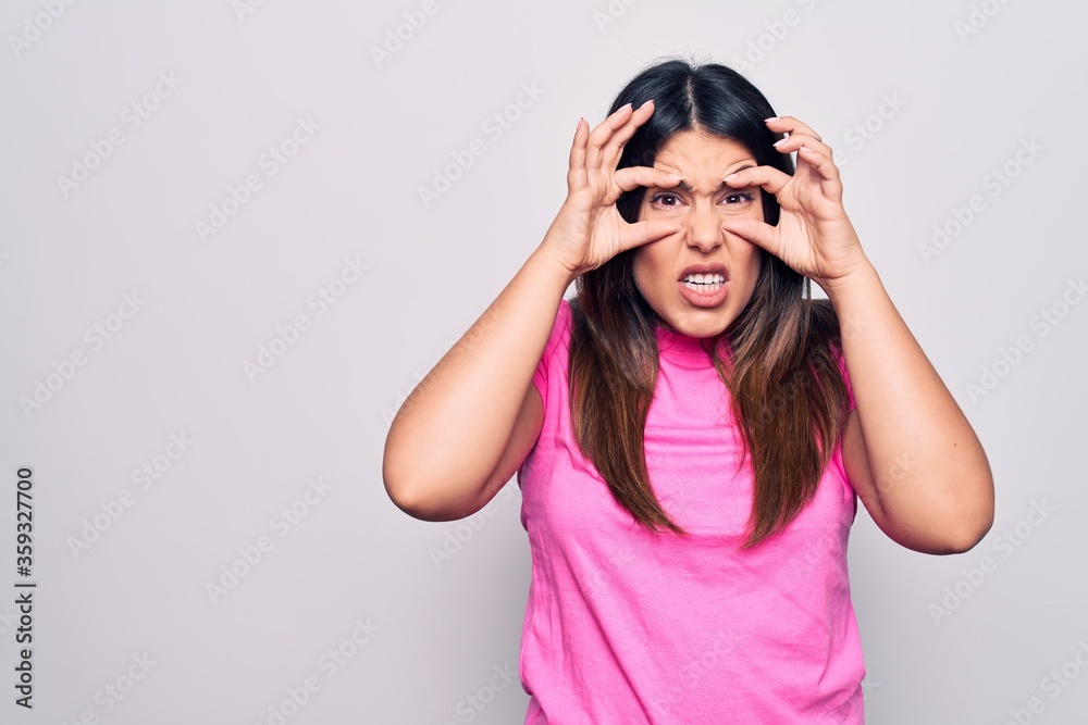 Young beautiful brunette woman wearing casual pink t-shirt standing over white background Trying to open eyes with fingers, sleepy and tired for morning fatigue Young beautiful brunette woman wearing casual pink t-shirt standing over white background Trying to open eyes with fingers, sleepy and tired for morning fatigue