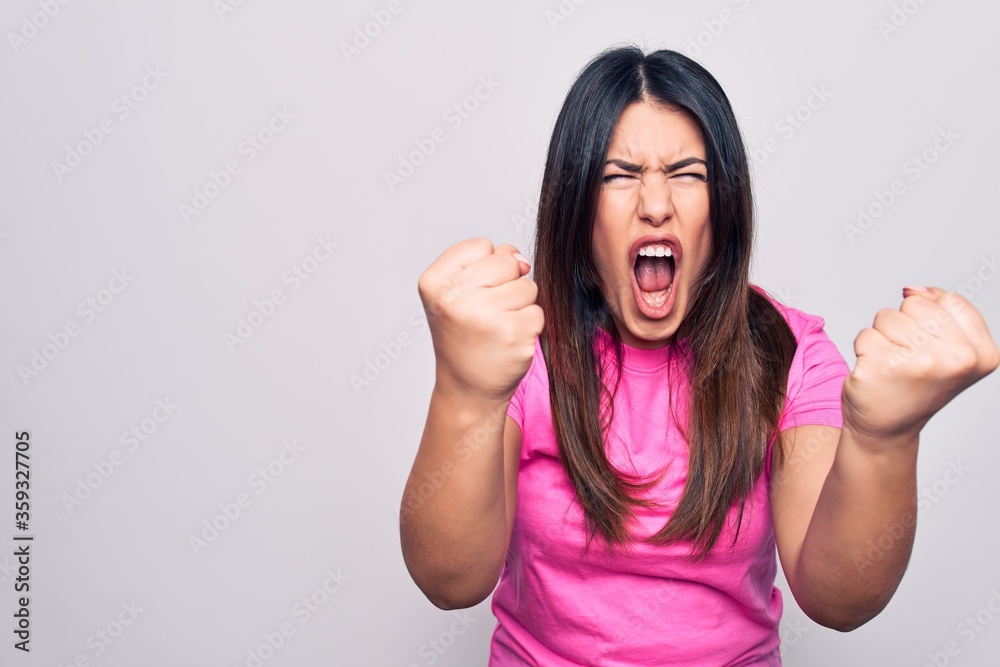 Young beautiful brunette woman wearing casual pink t-shirt standing over white background angry and mad raising fists frustrated and furious while shouting with anger. Rage and aggressive concept. Young beautiful brunette woman wearing casual pink t-shirt standing over white background angry and mad raising fists frustrated and furious while shouting with anger. Rage and aggressive concept.