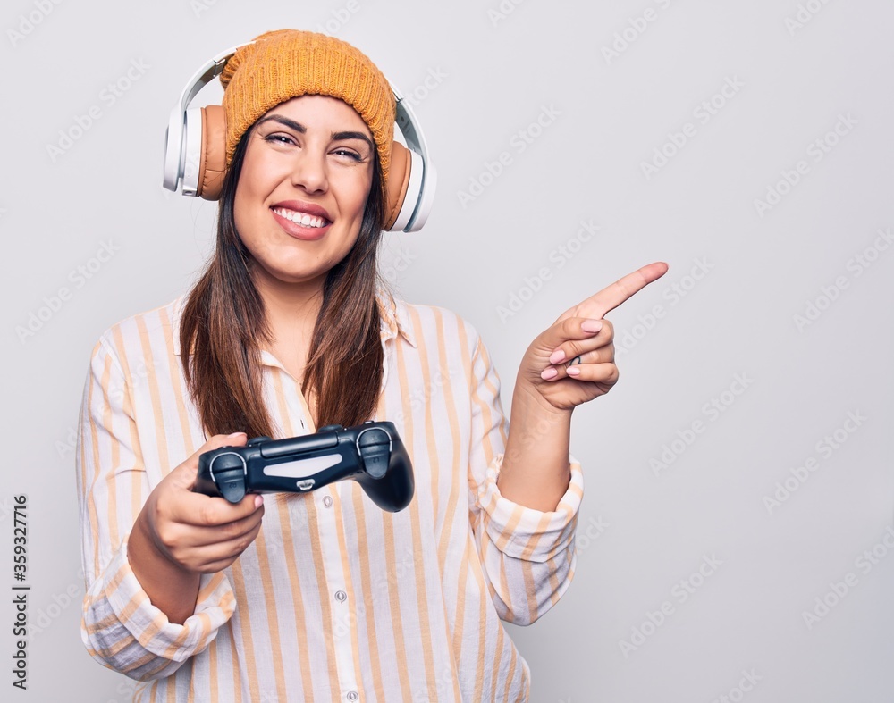 Young beautiful brunette gamer woman playing video game using joystick and headphones smiling happy pointing with hand and finger to the side Young beautiful brunette gamer woman playing video game using joystick and headphones smiling happy pointing with hand and finger to the side
