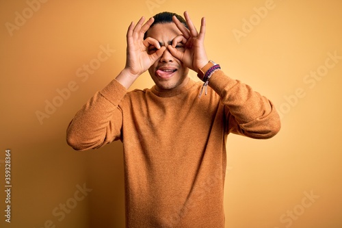 Young brazilian man wearing casual sweater standing over isolated yellow background doing ok gesture like binoculars sticking tongue out, eyes looking through fingers. Crazy expression.