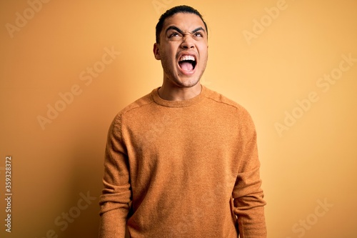 Young brazilian man wearing casual sweater standing over isolated yellow background angry and mad screaming frustrated and furious, shouting with anger. Rage and aggressive concept. © Krakenimages.com