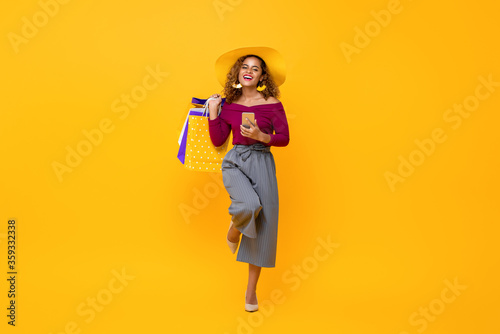Full length portrait of happy young attractive African American woman holding shopping bags and mobile phone with one leg raised in isolated studio yellow background