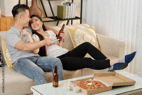 Young Vietnamese couple in love drinking beer and eating pizza when relaxing at home on Friday evening