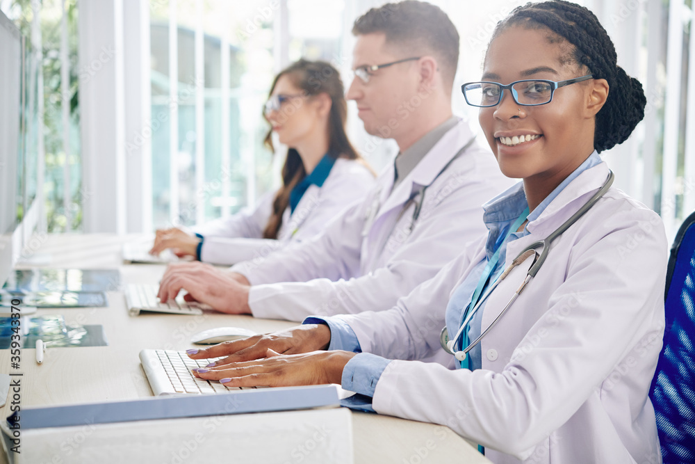 Fototapeta premium Smiling young Black medical researcher in goggles working on computer, her colleagues checking tests results near by