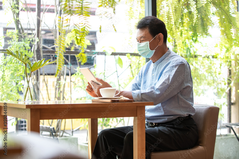 Old man using digital tablet in cafe and wearing mask for prevent the spread of the Covid-19, Covid-19 virus protection concept in Thailand.