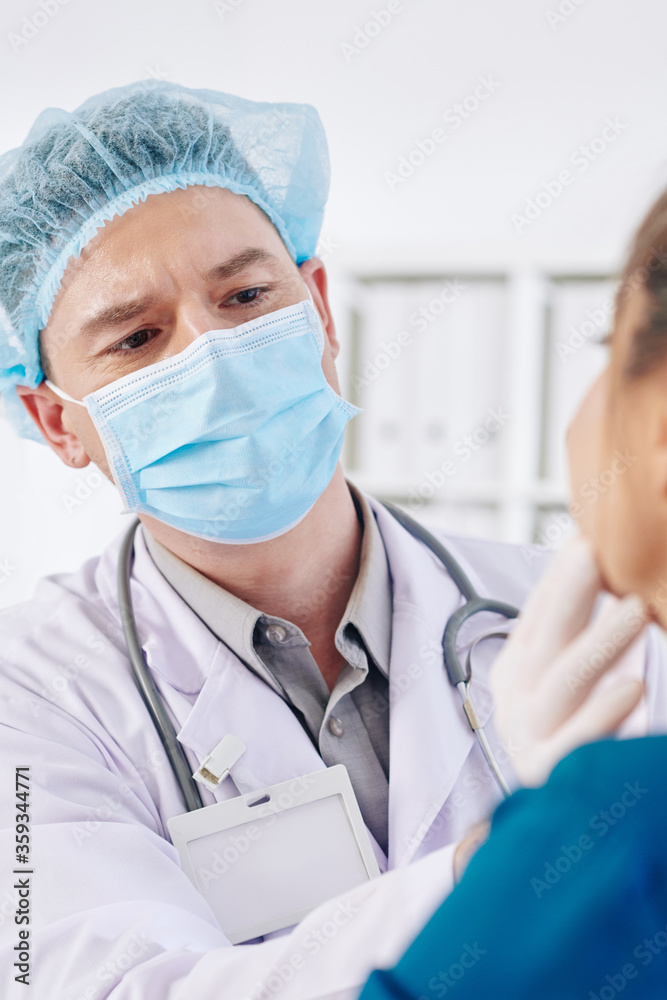 Serious general practitioner in medical mask examining glands and throat of female patient Serious general practitioner in medical mask examining glands and throat of female patient