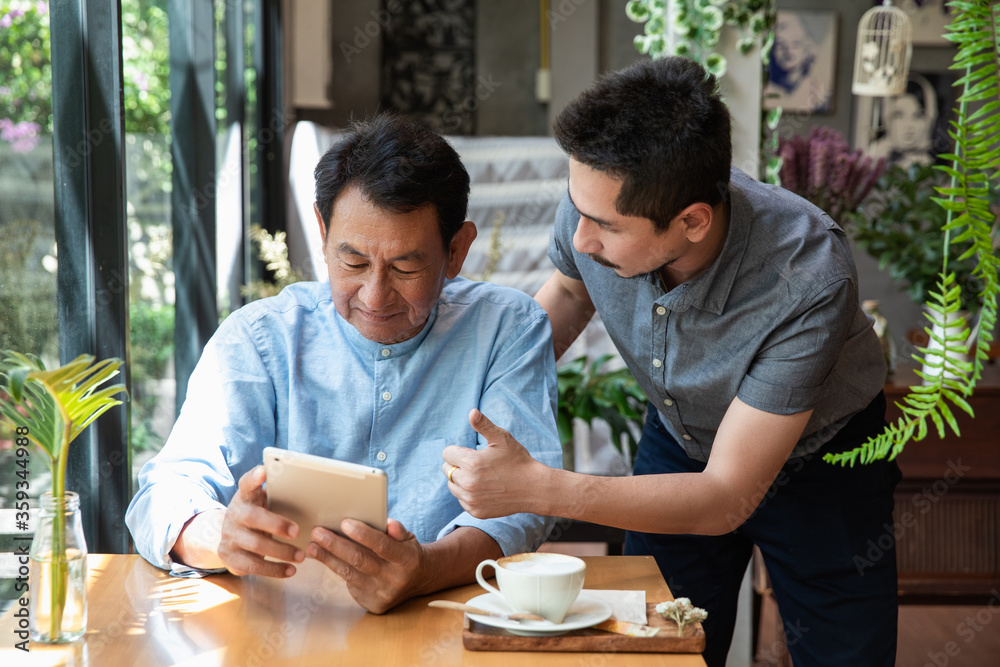 Portrait of a young man teaching his father how to use tablet computer. Son helping Dad to use the tablet until succeed and thumb up dad. Father and Son using a tablet together at cafe. Portrait of a young man teaching his father how to use tablet computer. Son helping Dad to use the tablet until succeed and thumb up dad. Father and Son using a tablet together at cafe.