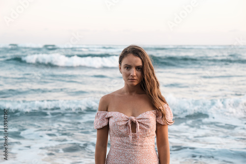 A sad beautiful young girl in a dress stands on the ocean and looks at the camera. Pleasant pastel colors. Girl close-up on a background of waves. The woman turned her back to the sea