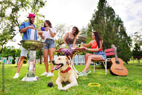 the Labrador Retriever dog is lying on the grass. A group or group of friends relax in nature  roast meat on the barbecue  play the guitar and smile against the background of greenery