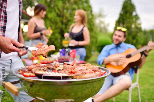 a man's hand holds a barbecue tongs with a juicy delicious meat steak against the background of a barbecue grill with meat and vegetables and a group of friends on a picnic who are having fun