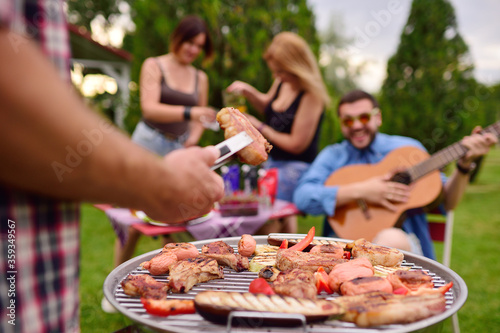 a man s hand holds a barbecue tongs with a juicy delicious meat steak against the background of a barbecue grill with meat and vegetables and a group of friends on a picnic who are having fun