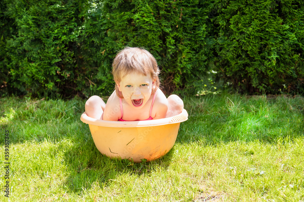 custom made wallpaper toronto digitalA little kid bathes in an orange basin in the summer in hot weather against a background of green bushes and grass. Girl looking at the camera, opened her mouth, emotions. The baby has blonde hair, 