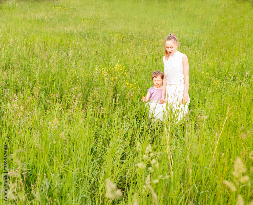 two little sisters hold hands, stand in a field. Around tall green grass, wildflowers. Girls examine blades of grass and little bugs. The summer warm sun warms them