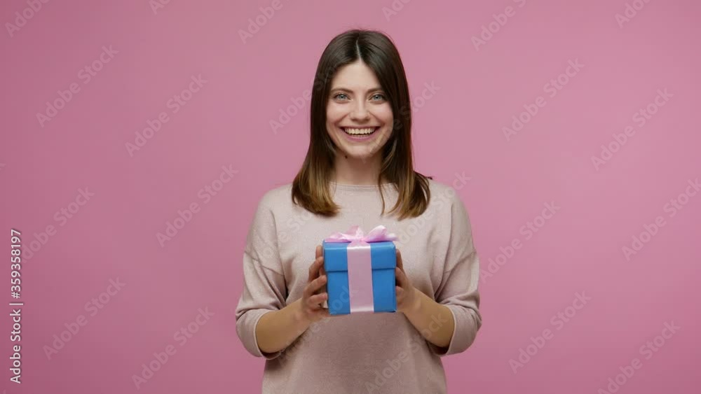 Charming woman catching wrapped present and giving box to camera, sharing christmas gift smiling friendly, celebrating holidays, congratulations on birthday. studio shot isolated on pink background