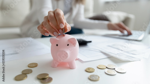 Close up coffee table lot of invoice, financial stats documents, utility bills, household payments, focus on female hand puts coin in piggy bank. Family budget manage, making personal savings concept