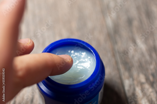 A closeup view of a finger dipping into a jar of vapor rub cream, on a wooden table surface.