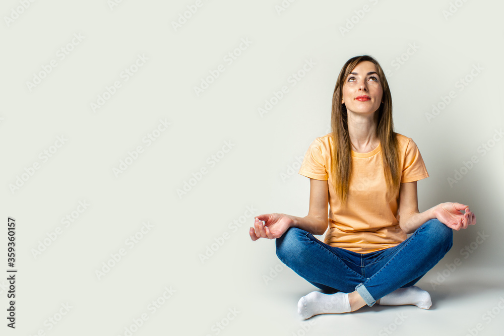 Young woman looks up in casual clothes and meditates while sitting on the floor on a light background. Relaxation Young woman looks up in casual clothes and meditates while sitting on the floor on a light background. Relaxation