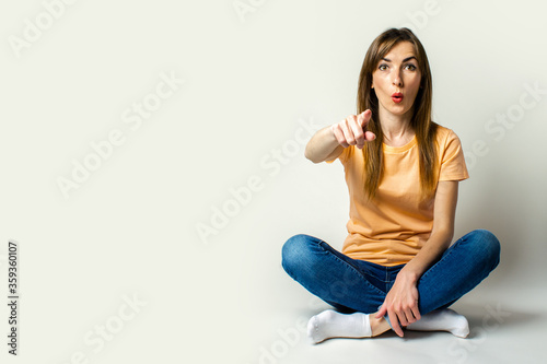 Young woman with a surprised face sits on the floor and points a finger at the viewer on a light background
