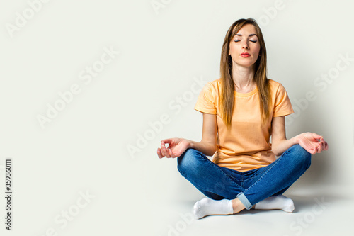 Young woman with closed eyes in casual wear meditates sitting on the floor on a light background