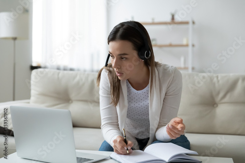 Focused businesswoman wear headphones writing in personal organizer during distant online communication with client using pc sit on couch at home. E-learn, language exam preparation, watching webinar