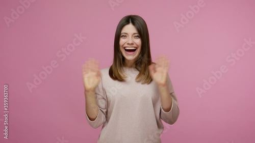 Lovely amiable brunette young woman waving hand to camera, greeting with hello gesture and smiling, welcoming saying hi with friendly expression. indoor studio shot isolated on pink background