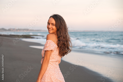 A beautiful young tourist girl walks along an empty black sand beach by the ocean in Bali. The girl in the dress smiles and turns around. Laughing smiling girl alone on the beach. 