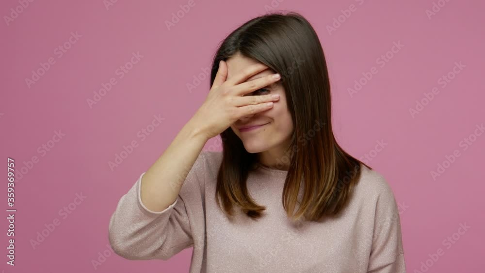 Funny playful brunette woman peeking through fingers, covering eyes with hand and smiling, curious about secret, shy to watch forbidden event, spying. indoor studio shot isolated on pink background