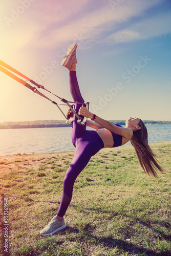 Fototapeta Naklejka Na Ścianę i Meble -  Young woman exercising with suspension trainer sling in park, near the lake.