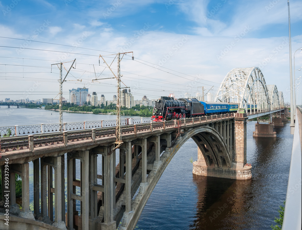 Passenger train with steam locomotive moves on bridge across river