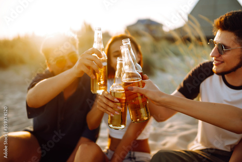 Young people sitting together at beach, drinking beer and having a party. Group of friends cheers with beers at the beach during summer vacation.