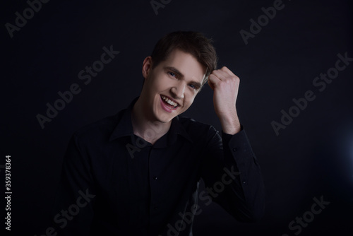 Studio portrait of a guy in a black shirt on a black background. A handsome young guy of European appearance. The guy laughs, the guy in a good mood looks at the camera