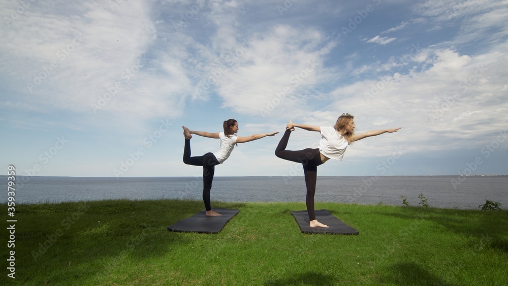 Two young women practice yoga on coast near the lake or sea. Training Lord of the Dance pose. Two young women practice yoga on coast near the lake or sea. Training Lord of the Dance pose.